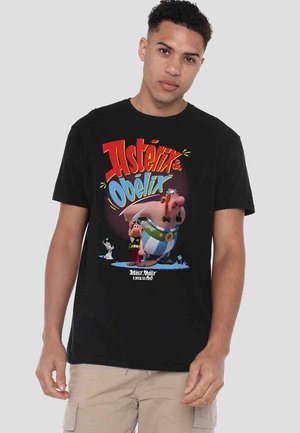 Man wearing black T-shirt with colorful Asterix and Obelix cartoon characters and logo, beige cargo pants, standing against plain background.