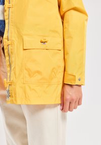 Veste imperméable jaune avec fermeture éclair, poche avant, accents de boutons et poignets ajustables. Présente une texture lisse et un design pratique.