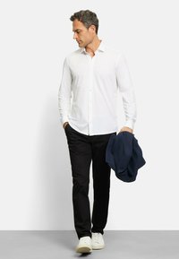 Weißes Hemd mit Knopfleiste, schwarze Hose und weiße Sneakers. Das Model hält eine dunkelblaue Jacke. Schlichtes Design, glatte Textur und maßgeschneiderte Passform.