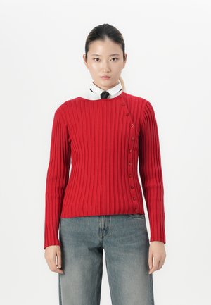Young woman with tied-back hair wears a red ribbed sweater buttoned diagonally over a white shirt and black tie, paired with faded jeans.