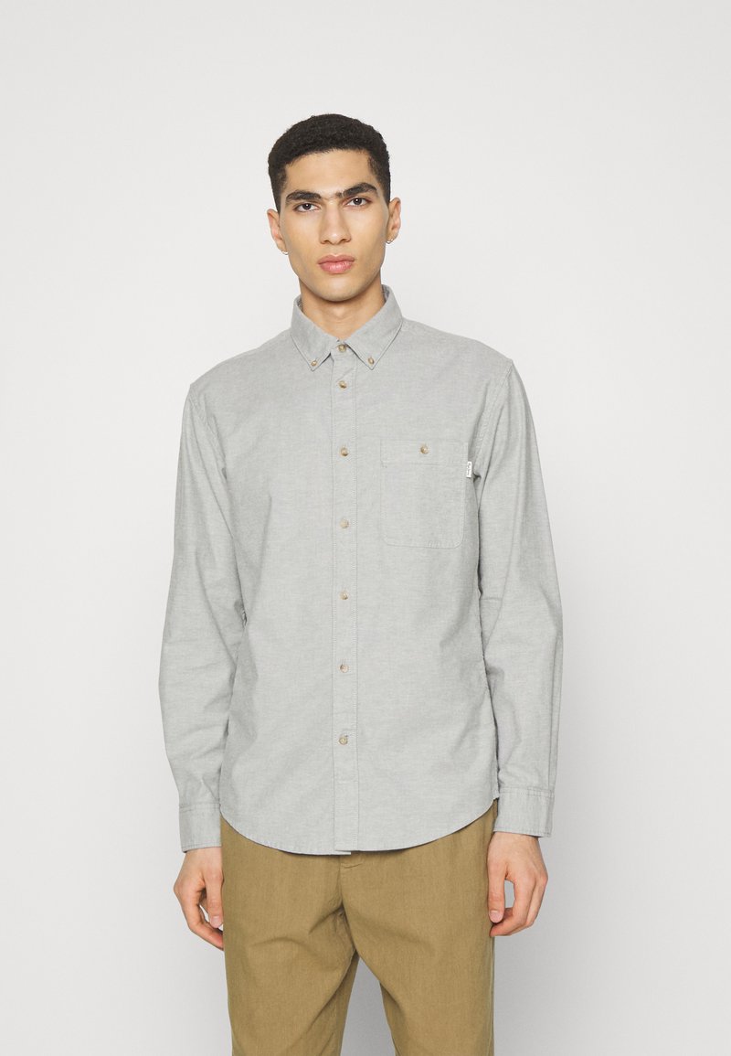 Camisa gris de manga larga con botones, con un bolsillo en el pecho, dobladillo redondeado y botones marrones. Llevada con pantalones de color marrón claro.