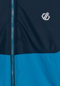 Blå och mörkblå vindjacka med dragkedja fram, utrustad med en liten vit logotyp på bröstet. Slät yta och lätt material.