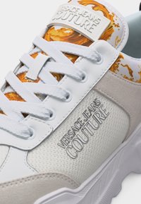 Λευκό και μπεζ αθλητικό παπούτσι Versace Jeans Couture με λευκά κορδόνια και πορτοκαλί μαρμάρινο μοτίβο στη μαξιλαρωμένη γλώσσα.