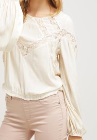 Långärmad krämfärgad blus med spetspaneler och rynkade manschetter, med en avslappnad passform och en något elastisk nederkant. Kombineras med ljusrosa byxor.