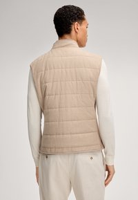 Beige, gesteppte Weste aus weichem Stoff, mit hohem Kragen und ärmellosem Design, getragen über einem weißen Langarmshirt.
