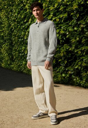 Grauer, gerippter Strick-Polo-Pullover mit Kragen, kombiniert mit weit geschnittenen, cremafarbenen Hosen und grauen Sneakers. Der Hintergrund zeigt grüne Blätter.
