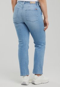 Gaiši zili džinsa bikses ar muguras kabatām, ko valkā cilvēks, kas stāv, apvienotas ar baltām sporta kurpēm un baltu topu.