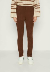 Person i brune slim-fit bukser, en kremfarget og brun stripet genser, og hvite sneakers med grønne striper som står mot en ensfarget bakgrunn.