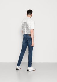 Camiseta blanca de manga corta con un estampado gráfico en la parte posterior, combinada con jeans ajustados de color azul y zapatillas blancas con detalles en negro.