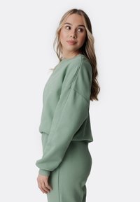 Felpa verde corta con colletto a costine e maniche ampie, realizzata in materiale morbido; abbinata a pantaloni coordinati. Design semplice con un branding sottile.