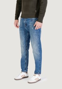Persona che indossa jeans blu sbiaditi, sneakers bianche con strisce beige e un maglione a coste verde scuro, in piedi su uno sfondo chiaro.