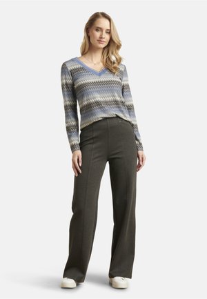 Woman with long blonde hair wearing a blue, gray, and white zigzag patterned sweater, dark gray wide-leg pants, and white sneakers.