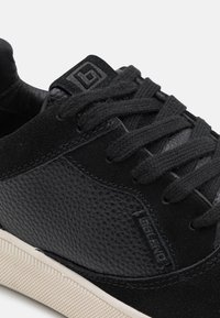 Black sneakers featuring a mix of textured suede and smooth leather, with flat black laces and a cream rubber sole. Close-up view.