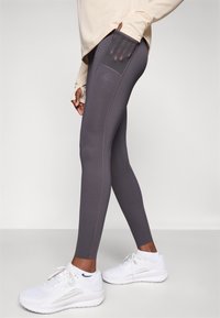 Grå leggings med en sidelomme, glatt tekstur og sømdetaljer, kombinert med hvite sportssko på en nøytral bakgrunn.
