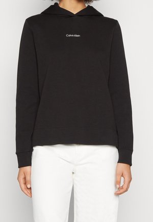Osoba nosząca czarną bluzę z kapturem Calvin Klein z białym logo na piersi i białe spodnie, stojąca na białym tle.