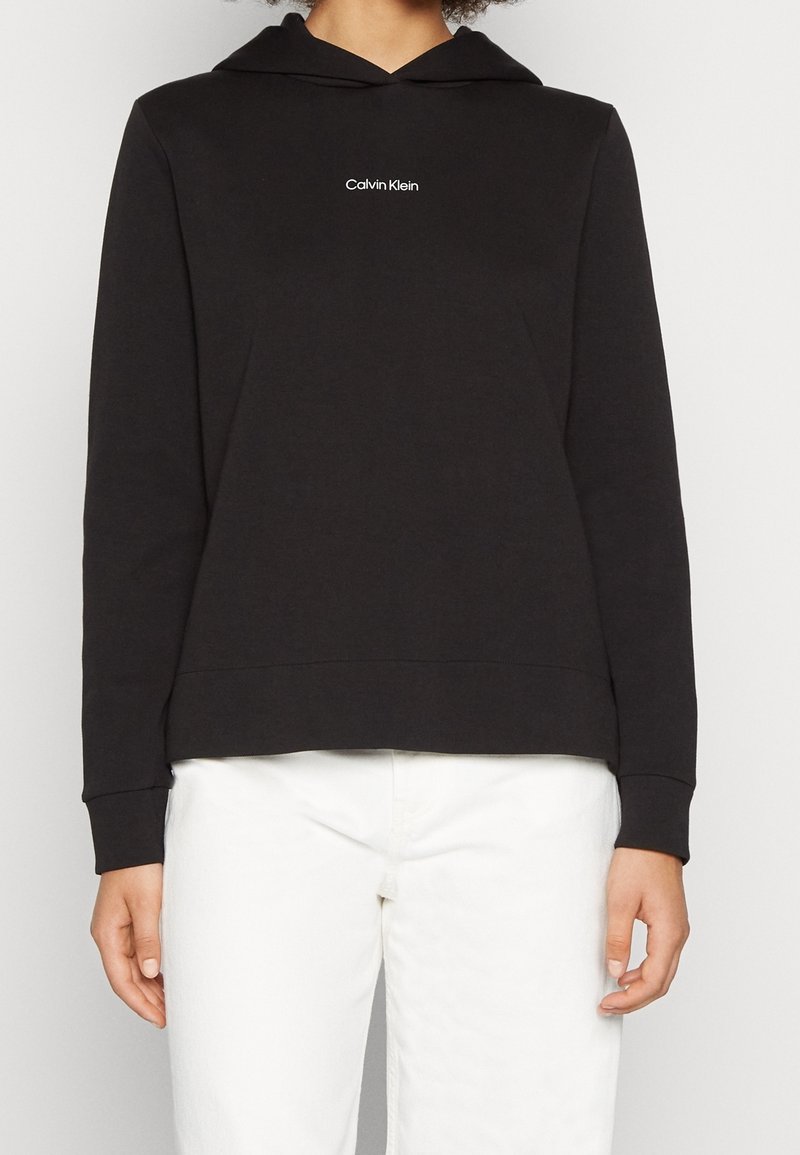 Personne portant un sweat à capuche noir Calvin Klein avec un logo blanc sur la poitrine et un pantalon blanc, debout devant un fond blanc.