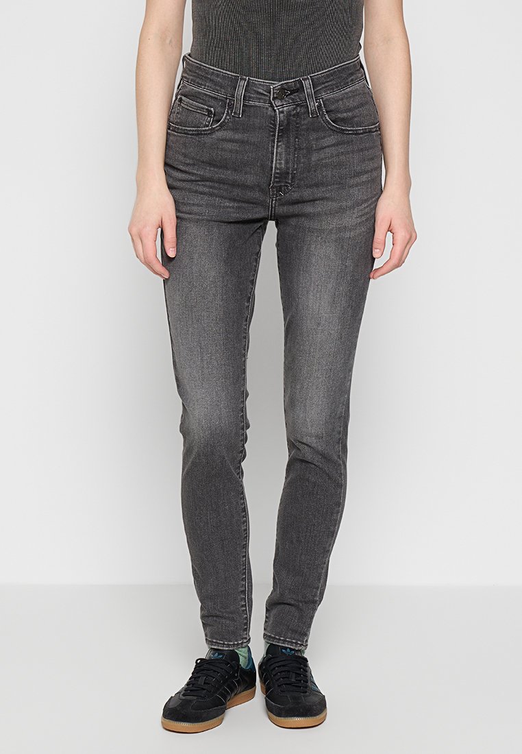Zwarte high-waisted skinny jeans met een gladde textuur, twee voorkruiden en een rits. Gecombineerd met zwarte sneakers en gemusterde sokken.