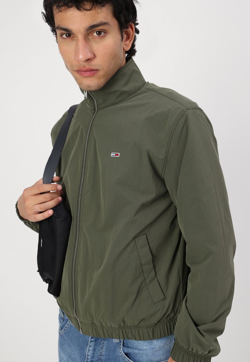 Veste zippée vert olive avec col montant, poignets élastiques, poches latérales et un petit patch logo. Associée à un jean bleu et un sac noir.