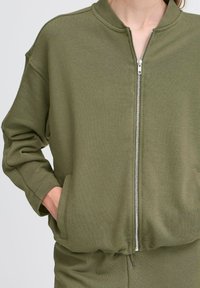 Olivovo zelená bundu na zips z jemnej látky s rebrovaným golierom, bočnými vreckami a mierne nadrozmernými rukávmi. Hladký, štruktúrovaný povrch.
