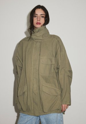 Kvinde med mørkt hår iført en oversized olivengrøn jakke med store lommer og lyseblå jeans, stående mod en ensfarvet baggrund.