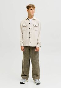 Ragazzo adolescente in piedi, con una giacca camicia beige a bottoni, pantaloni in velluto a coste marroni, sneaker bianche, sorridente e con lo sguardo leggermente a destra.