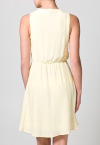 Vestido amarillo sin mangas con cintura ceñida, falda fluida y tela suave. La vista trasera muestra líneas limpias y sin adornos visibles.