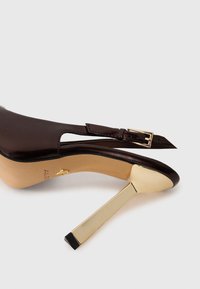 Ruske nahodne čevlje na visokem petu iz usnja z zlatim kovinskim stiletto peto in nastavljivim gležnjarjem z zlatim sponko, prikazani na beli ozadju.