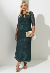 Kvinde i en mørkegrøn midi kjole med rynker og gennemsigtige 3/4 ærmer, som holder en gylden clutch og har gyldne sandaler og armbånd på.