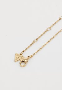 Collier en chaîne en or avec un pendentif triangulaire gravé du mot "GUESS" et un fermoir à ressort sécurisé.