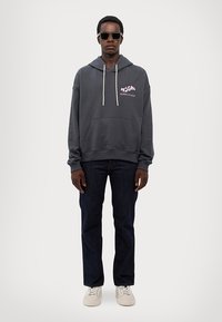 Grå hættetrøje med en frontlomme, hvide snore og pink tekstlogo. Båret med mørke jeans og hvide sneakers. Model iført solbriller.