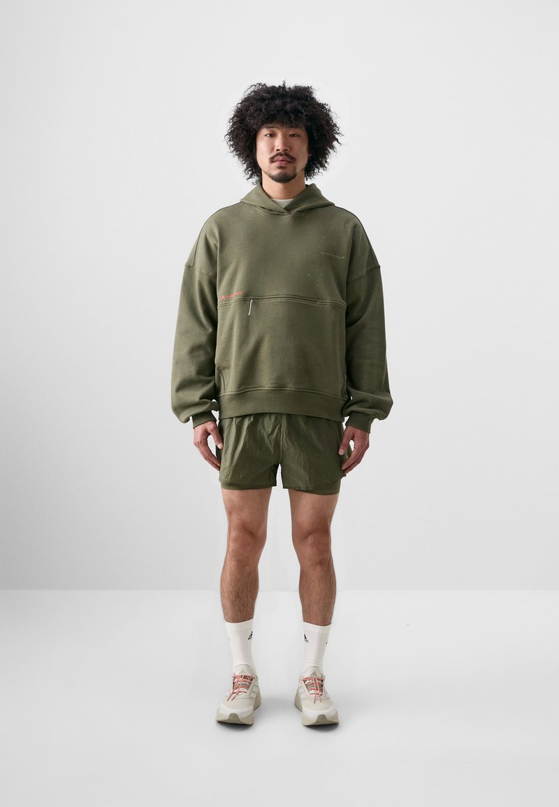 Olivengrøn oversized hoodie med frontlomme, parret med matchende letvægts shorts. Modellen har beige træningssko og hvide sokker på.