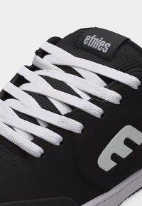 Μαύρο αθλητικό παπούτσι Etnies με λευκά κορδόνια, λευκό λογότυπο στο πλάι και ετικέτα με την επωνυμία στη γλώσσα, κοντινή λήψη στην περιοχή με τα κορδόνια.