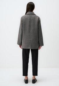 Cappotto grigio oversize in lana con maniche ampie, colletto a notched e dettaglio di cucitura sul retro, abbinato a pantaloni neri e scarpe.