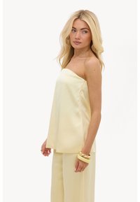 Femme blonde portant un top jaune clair sans bretelles et un pantalon large assorti, accessoirisée avec des bracelets dorés et jaunes.