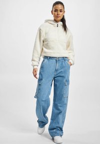 Hvit fleece zip-up hettejakke med foret hette, kombinert med lyseblå baggy cargo jeans med flere lommer. Hvite sneakers fullfører antrekket.