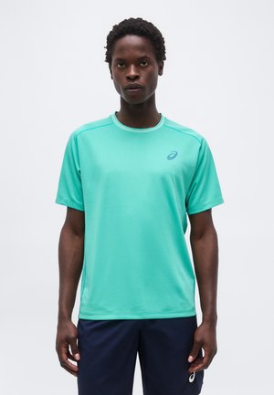 Άνδρας που φοράει τιρκουάζ αθλητική μπλούζα με κοντά μανίκια και σκούρο μπλε σορτς, στέκεται μπροστά σε απλό φωτεινό φόντο.