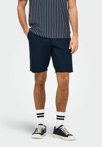Shorts en coton bleu marine à coupe décontractée, associés à une chemise à manches courtes rayée bleu marine et blanche et des baskets bleu marine avec semelles blanches et des chaussettes.