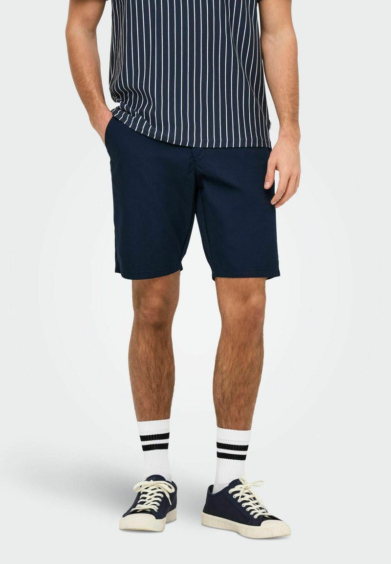 Shorts en coton bleu marine à coupe décontractée, associés à une chemise à manches courtes rayée bleu marine et blanche et des baskets bleu marine avec semelles blanches et des chaussettes.