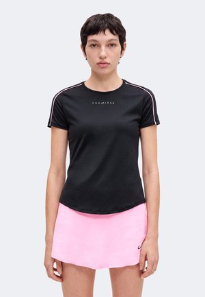Młoda kobieta z krótkimi ciemnymi włosami, ubrana w czarny t-shirt z napisem "ENDLESS" i jasnoróżową krótką spódnicę, stojąca na jednolitym tle.