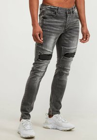 Vaqueros de mezclilla negros de corte ajustado con parches desgastados en las rodillas, acabado descolorido y adornos sutiles. Combinados con zapatillas deportivas blancas.