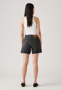Shorts de denim negro con dobladillos deshilachados, con un parche de cuero marrón en la parte trasera de la cinturilla y dos bolsillos traseros, mostrados desde atrás.