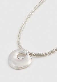 Harmaa kaulaketju, jossa on teksturoitu ketju ja epäsäännöllinen, sileä, donitsimuotoinen riipus, joka makaa litteänä valkoisella pinnalla.