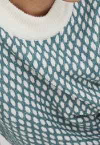 Pull en tricot avec une base turquoise et un motif en losanges blancs. Il comporte un col ras du cou côtelé blanc et une texture douce.