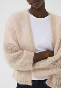 Lyserød, hyggelig cardigan lavet af fluffy mohair med løs pasform og ribbede manchetter, lagdelt over en hvid T-shirt. Struktureret stofdetalje synlig.