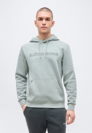 Giovane uomo con capelli corti che indossa una felpa con cappuccio Björn Borg grigio chiaro e pantaloni della tuta scuri, in piedi davanti a uno sfondo bianco uniforme.