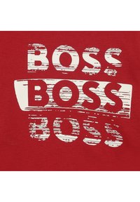 Sarkans T-krekls ar lieliem, nolietotiem baltiem burtiem "BOSS" dažādās izmērās, ar otas trieciena dizainu un gludu auduma tekstūru.