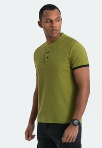 Polo shirt verde a maniche corte con una finitura testurizzata, colletto con tre bottoni e dettagli neri sulle maniche. Orologio nero al polso.