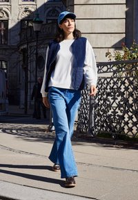 Femme marchant à l'extérieur par une journée ensoleillée, portant une casquette bleue, un gilet bleu marine, une chemise blanche à manches longues, un jean bleu et des chaussures marron près d'une clôture métallique ornée.
