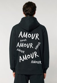 Čierna mikina s kapucňou, ktorá na chrbte obsahuje viacero bielych nápisov "AMOUR" rôznych veľkostí. Mäkká látka s voľným strihom a kapucňou.