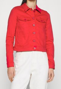 Chaqueta de mezclilla roja recortada con cuello, cierre de botones en el frente, dos bolsillos en el pecho y dos bolsillos laterales, con botones rosas como acentos.